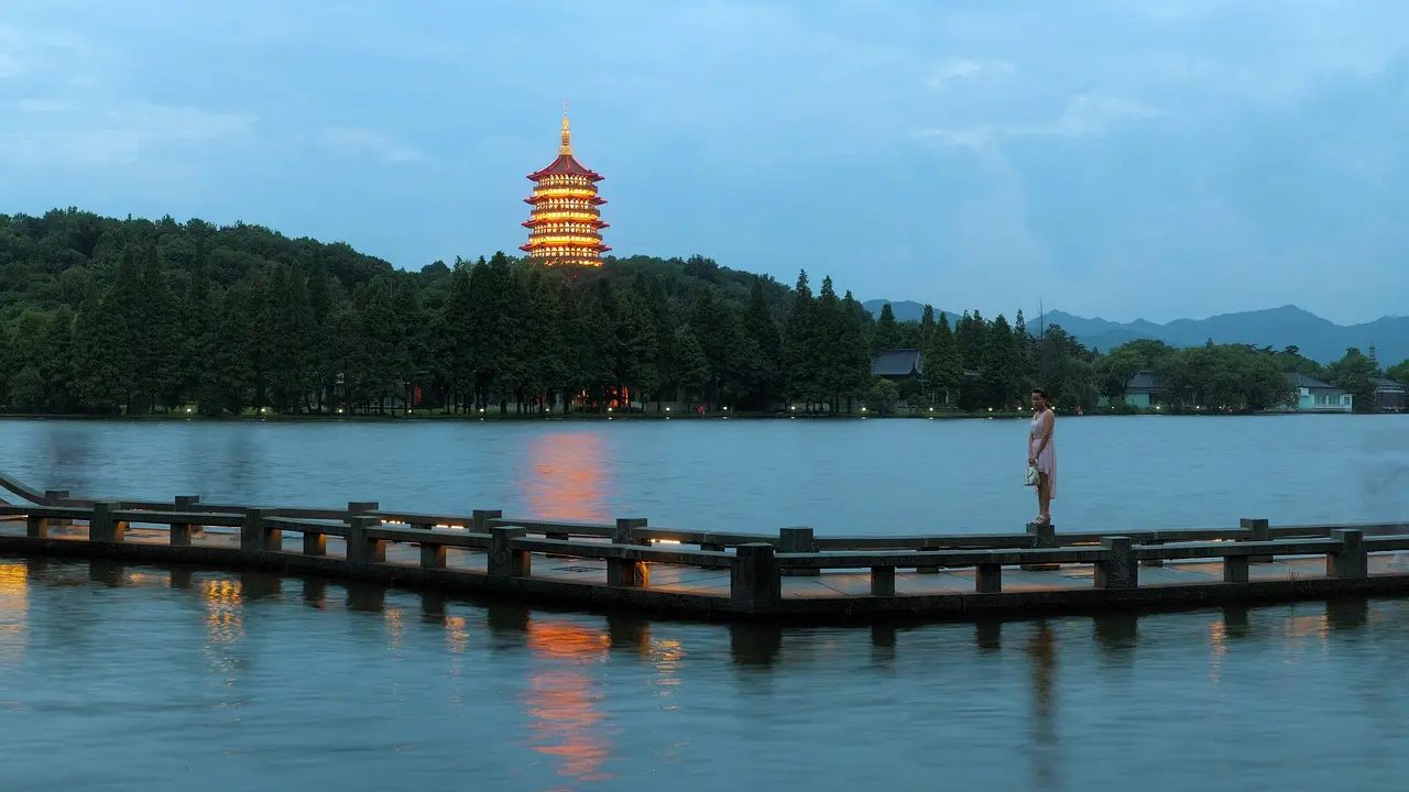 Barco tradicional no Lago Oeste de Hangzhou, com pagode e montanhas ao fundo sob céu nublado suave.