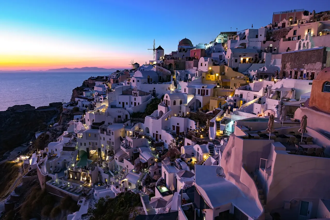 Vista aérea de casas brancas e cúpulas azuis em Santorini ao entardecer, com o mar ao fundo.